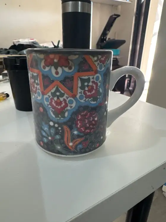Seller's phone photo: a ceramic mug with a full-wrap floral pattern on a cluttered work table