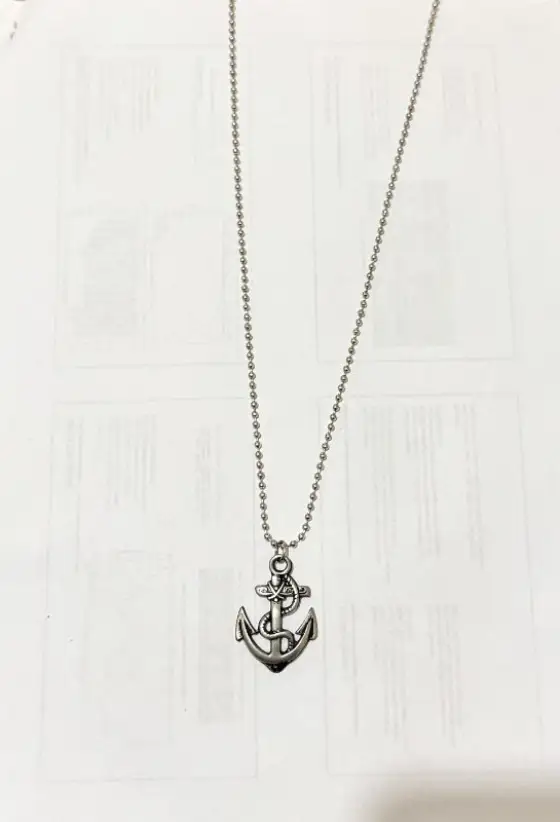 Seller's phone photo: a silver-tone anchor pendant on a ball chain, laid on printed paper