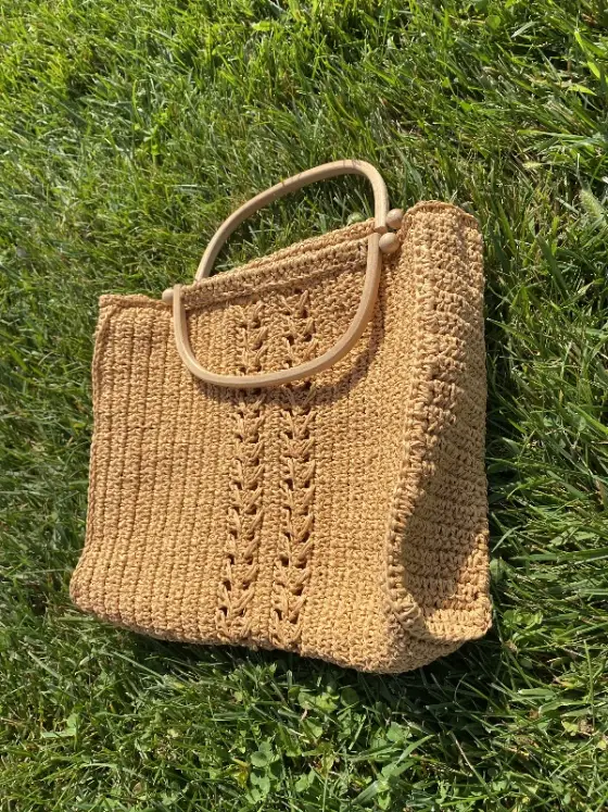 Seller's phone photo: a straw-coloured crochet tote bag with round wooden handles, lying on grass